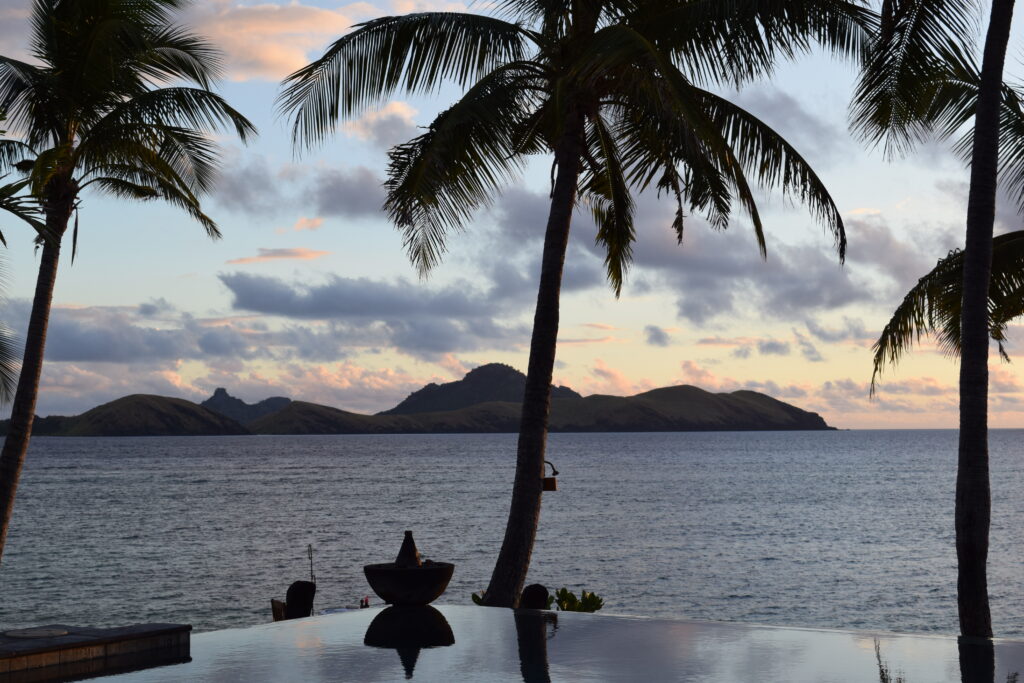 Sunset over a swim pool in Fiji. Framed by multiple palm trees and an island in the distance.