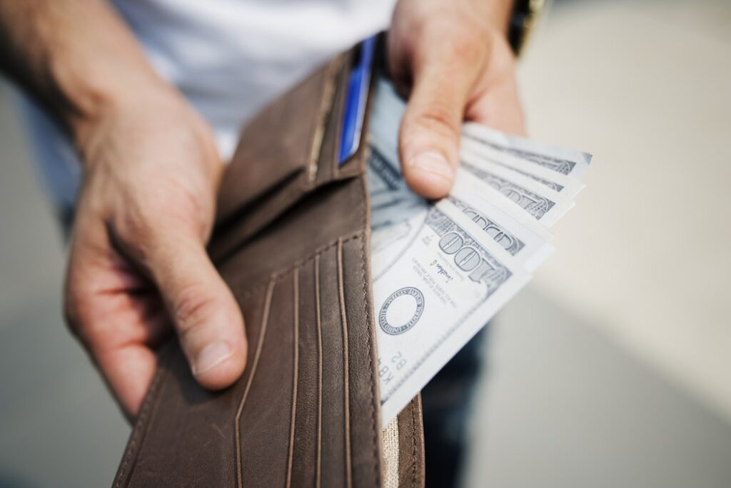 A man's hands holding an open wallet, with partially pulled out 100 dollar bills showing money saved on hotels.